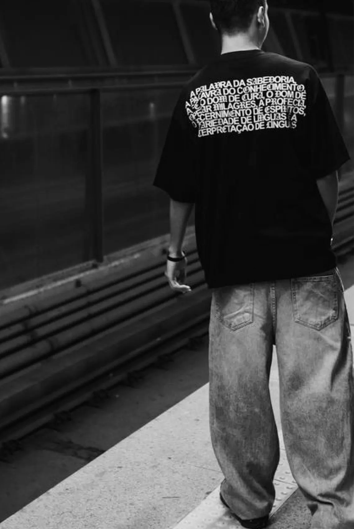 A model in an oversized black YAWI T-shirt on a station platform, the green and white hand tags at his side.