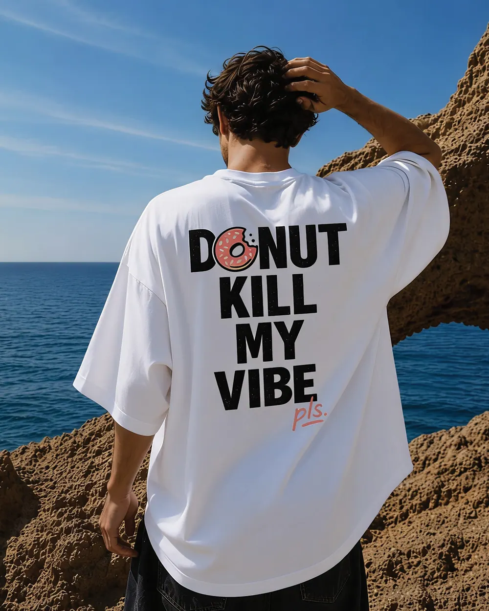 The back of a white YAWI tee printed with the Donut kill my vibe graphic, worn by the sea.