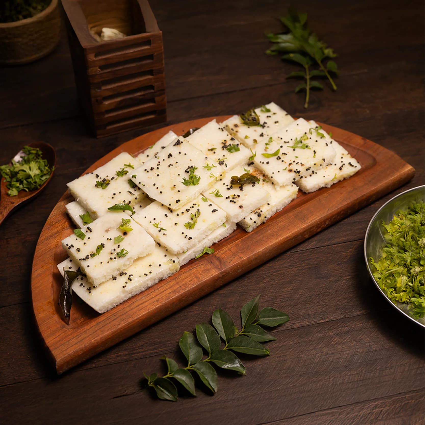 A food photograph of idada: white squares topped with mustard seeds, curry leaves and coriander, on a wooden board.